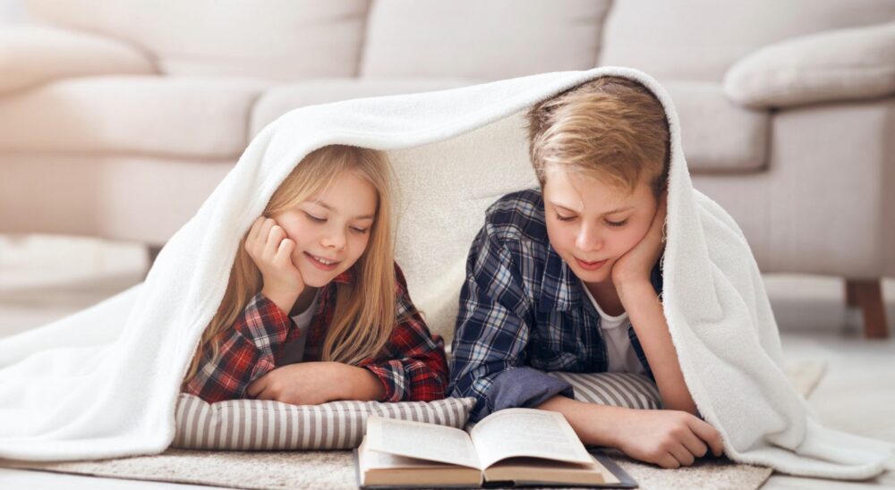 Dos niños tumbados en el suelo bajo una manta blanca, leyendo un libro juntos
