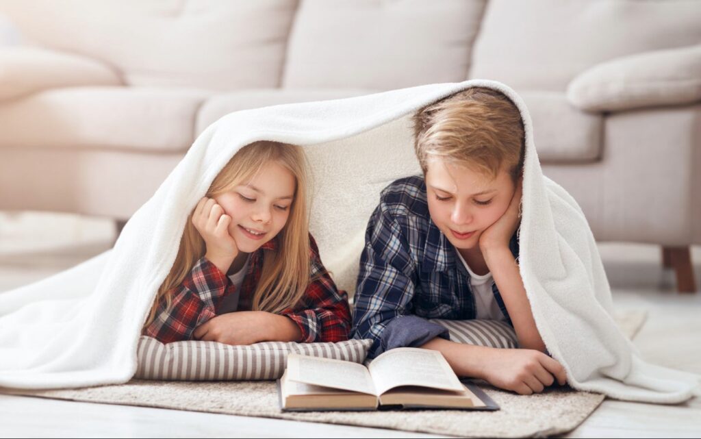 Dos niños tumbados en el suelo bajo una manta blanca, leyendo un libro juntos