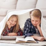 Dos niños tumbados en el suelo bajo una manta blanca, leyendo un libro juntos